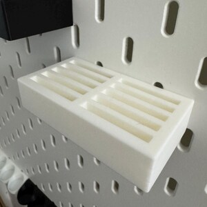 Op de afbeelding: Een witte pegboard plank met meerdere sleuven voor opslag. Een zwarte pegboard plank is ook zichtbaar. De planken zijn ontworpen om te worden bevestigd aan een witte pegboard met ovale gaten.