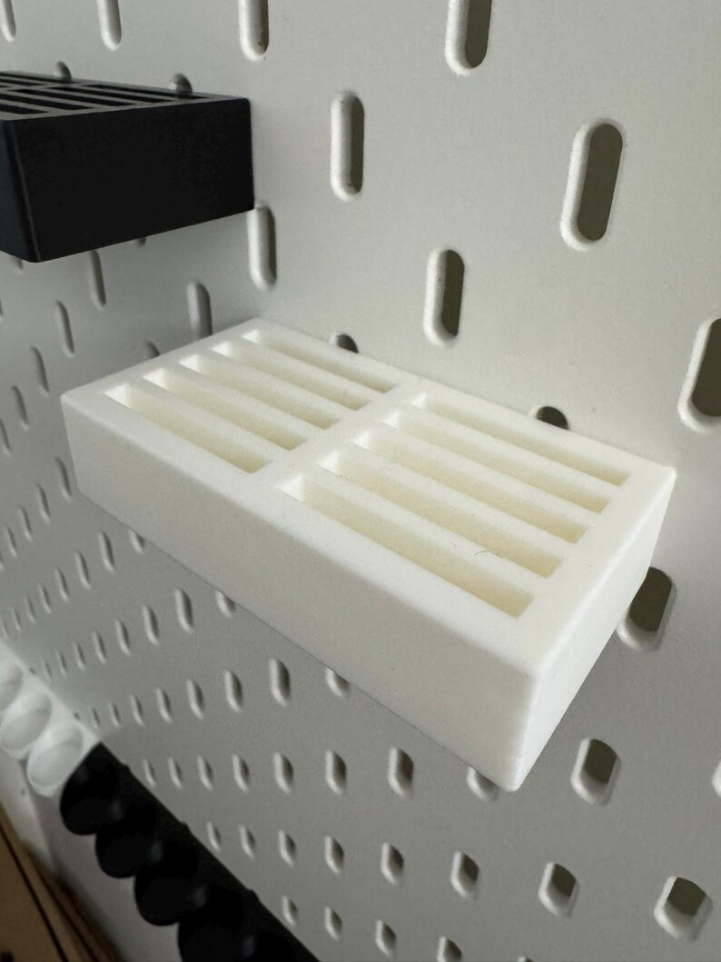 Op de afbeelding: Een witte pegboard plank met meerdere sleuven voor opslag. Een zwarte pegboard plank is ook zichtbaar. De planken zijn ontworpen om te worden bevestigd aan een witte pegboard met ovale gaten.
