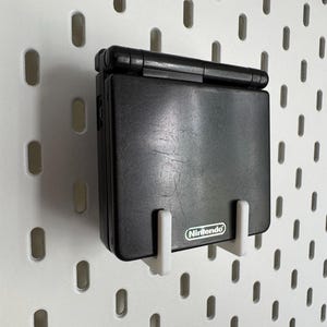 May include: A black Nintendo Game Boy Advance SP handheld console mounted on a white pegboard. The console is held in place by two white brackets. The Nintendo logo is visible on the console. The pegboard has a series of oval-shaped holes.