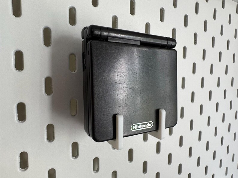 May include: A black Nintendo Game Boy Advance SP handheld console mounted on a white pegboard. The console is held in place by two white brackets. The Nintendo logo is visible on the console. The pegboard has a series of oval-shaped holes.