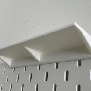 Puede incluir: Estante de pl&aacute;stico blanco con dos compartimentos, montado en una pared de tablero de clavijas blanco.