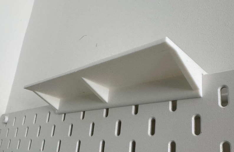 Puede incluir: Estante de pl&aacute;stico blanco con dos compartimentos, montado en una pared de tablero de clavijas blanco.
