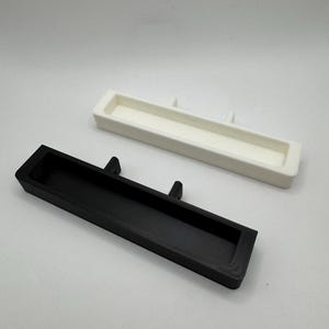 May include: Two rectangular, open-top trays. One is black, the other is white. Each tray has two small, raised tabs on one of the long sides. The trays are likely for organizing small items.