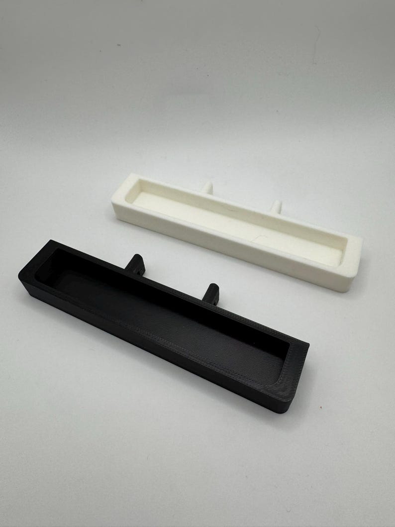 May include: Two rectangular, open-top trays. One is black, the other is white. Each tray has two small, raised tabs on one of the long sides. The trays are likely for organizing small items.