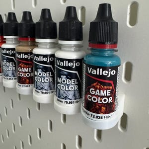以下が含まれることがあります： 黒いキャップと白いラベルが付いた、Vallejo Model ColorとGame Colorのペイントボトルの列。ボトルは白いペグボードに表示されています。ラベルにはブランド名と色の情報が含まれています。色は白、茶色、ターコイズブルーなどです。