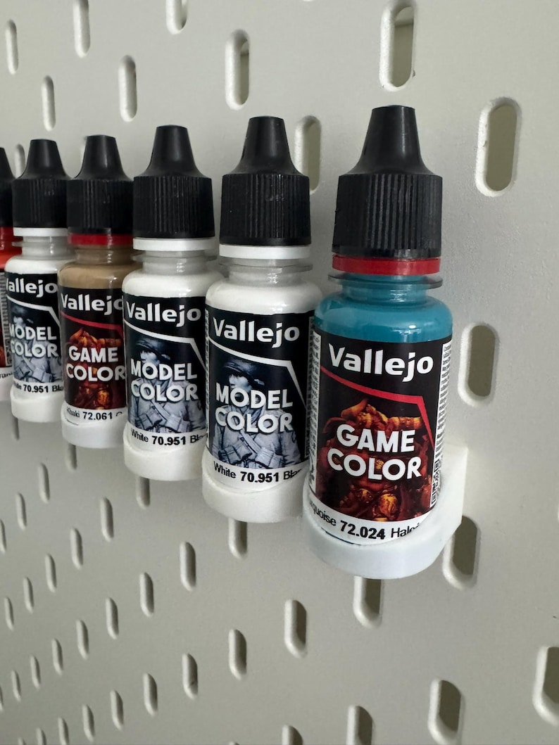 以下が含まれることがあります： 黒いキャップと白いラベルが付いた、Vallejo Model ColorとGame Colorのペイントボトルの列。ボトルは白いペグボードに表示されています。ラベルにはブランド名と色の情報が含まれています。色は白、茶色、ターコイズブルーなどです。