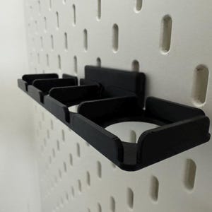 K&ouml;nnte beinhalten: Ein schwarzer, 3D-gedruckter Lochwand-Organizer mit mehreren F&auml;chern. Der Organizer ist an einer wei&szlig;en Lochwand mit gleichm&auml;&szlig;ig verteilten L&ouml;chern befestigt. Das Design umfasst Schlitze und einen kreisf&ouml;rmigen Halter, ideal zum Organisieren kleiner Gegenst&auml;nde.