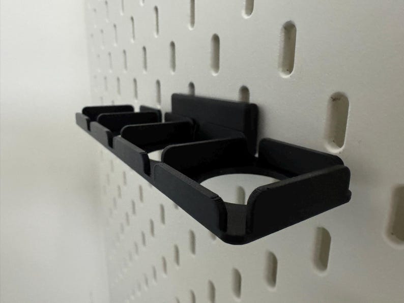 K&ouml;nnte beinhalten: Ein schwarzer, 3D-gedruckter Lochwand-Organizer mit mehreren F&auml;chern. Der Organizer ist an einer wei&szlig;en Lochwand mit gleichm&auml;&szlig;ig verteilten L&ouml;chern befestigt. Das Design umfasst Schlitze und einen kreisf&ouml;rmigen Halter, ideal zum Organisieren kleiner Gegenst&auml;nde.