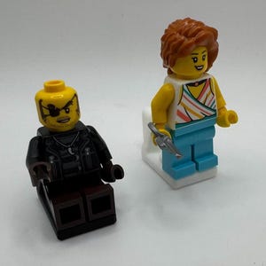 K&ouml;nnte beinhalten: Zwei Lego-Minifiguren. Eine Figur hat einen gelben Kopf mit Augenklappe und eine schwarze Jacke. Die andere Figur hat einen gelben Kopf, orangefarbene Haare, ein gestreiftes Oberteil, blaue Hosen und h&auml;lt eine silberne Pistole.