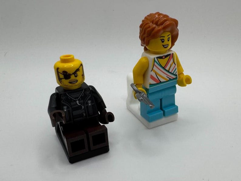 K&ouml;nnte beinhalten: Zwei Lego-Minifiguren. Eine Figur hat einen gelben Kopf mit Augenklappe und eine schwarze Jacke. Die andere Figur hat einen gelben Kopf, orangefarbene Haare, ein gestreiftes Oberteil, blaue Hosen und h&auml;lt eine silberne Pistole.