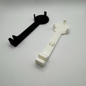 K&ouml;nnte beinhalten: Zwei 3D-gedruckte Handyst&auml;nder, einer schwarz und einer wei&szlig;. Beide St&auml;nder haben eine runde Basis mit einem erh&ouml;hten Rand und einen gebogenen Arm, der sich nach au&szlig;en erstreckt, um ein Handy zu halten.