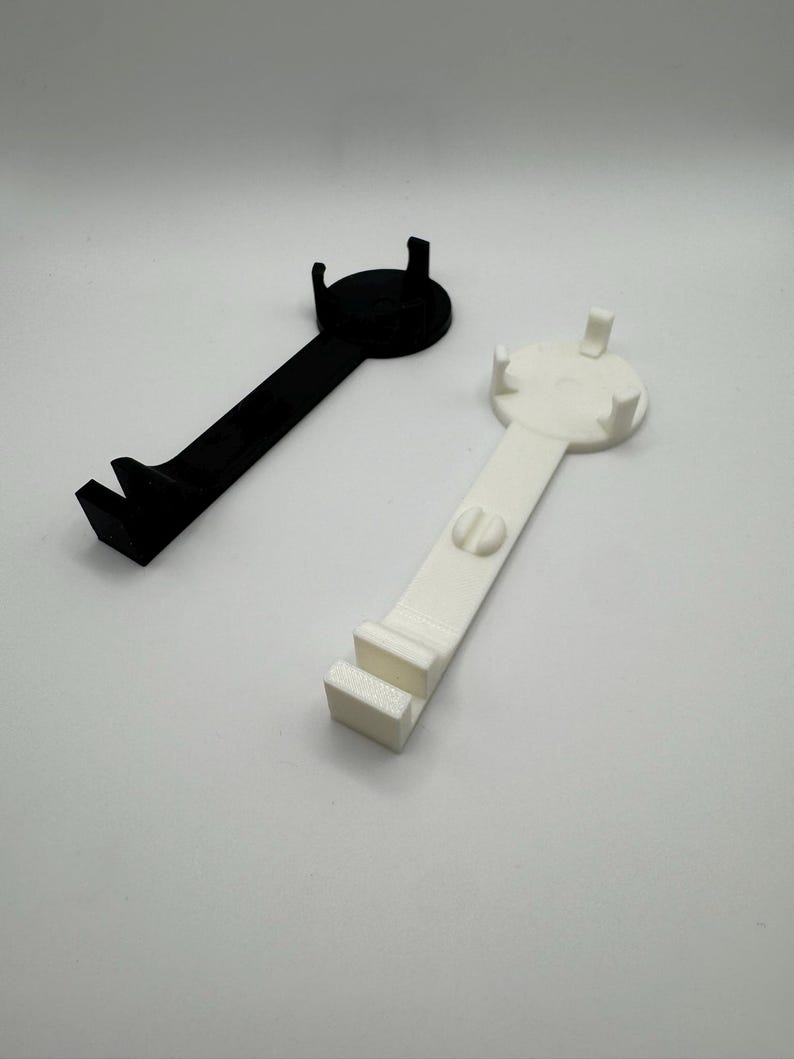 K&ouml;nnte beinhalten: Zwei 3D-gedruckte Handyst&auml;nder, einer schwarz und einer wei&szlig;. Beide St&auml;nder haben eine runde Basis mit einem erh&ouml;hten Rand und einen gebogenen Arm, der sich nach au&szlig;en erstreckt, um ein Handy zu halten.