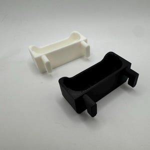 以下が含まれることがあります： 2つの長方形の3Dプリントオブジェクト。1つは白、もう1つは黒です。それぞれが湾曲した上部と2つの突き出た長方形のサポートを持っています。オブジェクトは、おそらくより大きなアイテムのコンポーネントまたはパーツです。