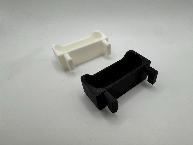 以下が含まれることがあります： 2つの長方形の3Dプリントオブジェクト。1つは白、もう1つは黒です。それぞれが湾曲した上部と2つの突き出た長方形のサポートを持っています。オブジェクトは、おそらくより大きなアイテムのコンポーネントまたはパーツです。