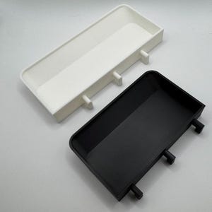 Puede incluir: Dos bandejas peque&ntilde;as rectangulares, una blanca y otra negra, con tres peque&ntilde;os pomos redondos en el lateral. Las bandejas est&aacute;n hechas de un material liso, similar al pl&aacute;stico.