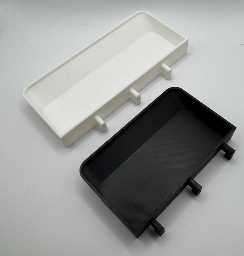 Puede incluir: Dos bandejas peque&ntilde;as rectangulares, una blanca y otra negra, con tres peque&ntilde;os pomos redondos en el lateral. Las bandejas est&aacute;n hechas de un material liso, similar al pl&aacute;stico.