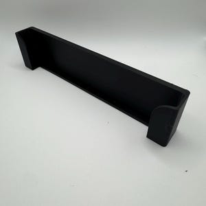 Op de afbeelding: Zwarte kunststof plankbeugel met een gebogen rand. De beugel is ontworpen om aan een muur of ander oppervlak te worden gemonteerd.