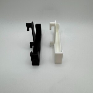 Puede incluir: Dos clips de pl&aacute;stico, uno negro y otro blanco, se muestran sobre un fondo blanco. Cada clip tiene un cuerpo rectangular con un gancho en la parte superior y otro m&aacute;s peque&ntilde;o en la parte inferior, dise&ntilde;ado para colgar.
