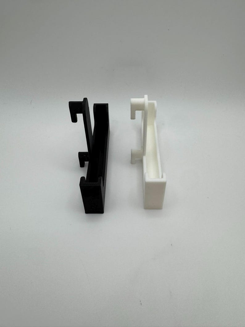Puede incluir: Dos clips de pl&aacute;stico, uno negro y otro blanco, se muestran sobre un fondo blanco. Cada clip tiene un cuerpo rectangular con un gancho en la parte superior y otro m&aacute;s peque&ntilde;o en la parte inferior, dise&ntilde;ado para colgar.