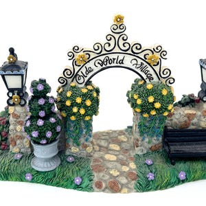 May include: A miniature Old World Village scene with a stone path leading to an archway inscribed with "Old World Village". The scene includes a black bench, two street lamps, and decorative shrubbery with purple and yellow flowers.