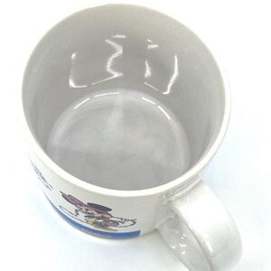 Op de afbeelding: Witte keramische mok met een cartoonafbeelding van Minnie Mouse. De mok heeft een gebogen handvat en een blauwe streep met tekst. De binnenkant van de mok is lichtgrijs van kleur. De mok is leeg en staat op een wit oppervlak.