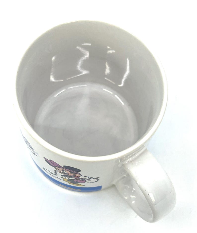 Op de afbeelding: Witte keramische mok met een cartoonafbeelding van Minnie Mouse. De mok heeft een gebogen handvat en een blauwe streep met tekst. De binnenkant van de mok is lichtgrijs van kleur. De mok is leeg en staat op een wit oppervlak.