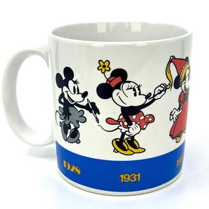 Op de afbeelding: Witte keramische mok met stripfiguren. De mok heeft een blauwe band aan de onderkant met het jaar 1931. De mok toont Minnie Mouse in een rode jurk en gele schoenen, en Mickey Mouse in zwart en wit.