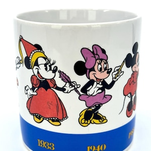 Op de afbeelding: Witte keramische mok met stripfiguren in verschillende outfits. De mok heeft een blauwe band aan de onderkant met de jaren 1933 en 1940 in goud. De stripfiguren zijn in rood, geel, zwart en paars.