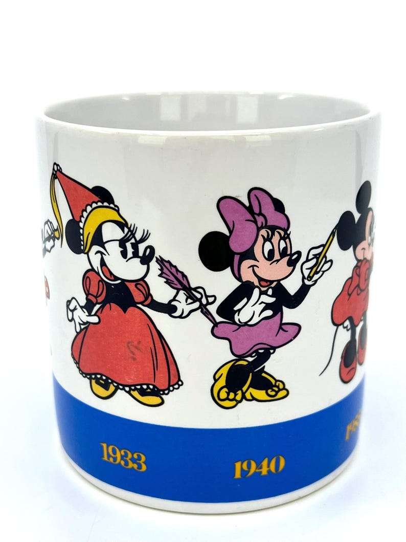 Op de afbeelding: Witte keramische mok met stripfiguren in verschillende outfits. De mok heeft een blauwe band aan de onderkant met de jaren 1933 en 1940 in goud. De stripfiguren zijn in rood, geel, zwart en paars.