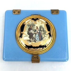 May include: A light blue compact mirror with a gold-toned hinged lid. The lid features a round, gold-rimmed design with a black and gold background and a painted scene of a couple in a garden.