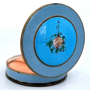 Op de afbeelding: Een blauwe en gouden emaille compact met een geschilderd bloemenontwerp. De compact is open en de binnenkant is lichtroze.