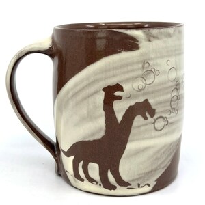 May include: A brown ceramic mug with a white glaze. The mug features a silhouette of a two-headed creature with bubbles around it.