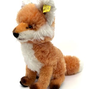 Puede incluir: Un peluche de zorro con pelaje naranja, blanco y marrón. El zorro tiene una nariz negra, ojos marrones y una cola tupida. Una etiqueta amarilla está adherida a su oreja con el texto "Covering 80% nylon 20% cotton."