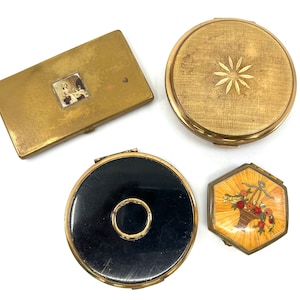 Op de afbeelding: Vier vintage compact spiegels. Eén is goud met een bloemenontwerp, één is goud met een zwart midden, één is goud met een stervormig ontwerp en één is goud met een klein plaatje van een vrouw.