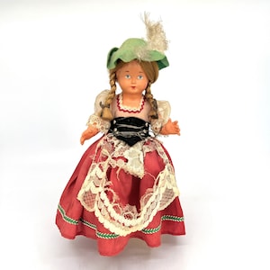 May include: A vintage doll wearing a red and white dress with lace trim, a green hat with a feather, and a black belt. The doll has blonde hair and blue eyes.