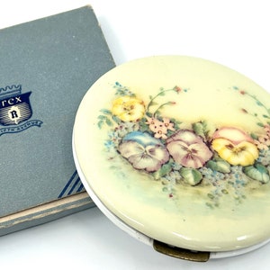 Op de afbeelding: Een vintage compact spiegel met een bloemenmotief. De spiegel is wit met een crèmekleurige achtergrond en heeft een geschilderd ontwerp van paarse, gele en roze viooltjes met groene bladeren en blauwe bloemen. De compact heeft een messing sluiting.