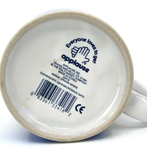 Op de afbeelding: Witte keramische mok met een oor, met de tekst "Everyone loves to get" en het Applause-logo. De onderkant van de mok bevat de tekst "Minnie Draws Minnie" en een barcode. Vaatwasser- en magnetronbestendig.