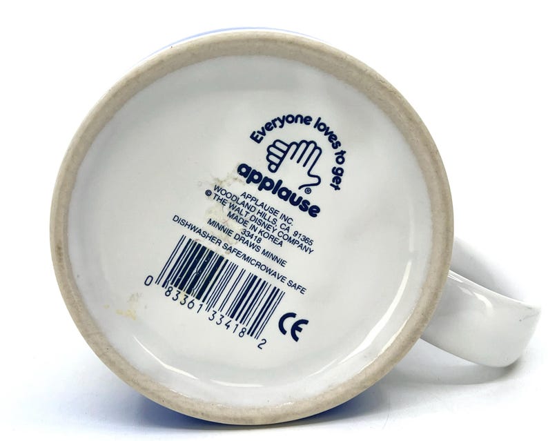 Op de afbeelding: Witte keramische mok met een oor, met de tekst "Everyone loves to get" en het Applause-logo. De onderkant van de mok bevat de tekst "Minnie Draws Minnie" en een barcode. Vaatwasser- en magnetronbestendig.