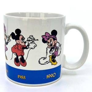 Op de afbeelding: Witte keramische mok met Minnie Mouse in verschillende poses. De mok heeft een blauwe band aan de onderkant met de jaren 1955 en 1990 in goud. De handgreep is wit en de mok is cilindrisch.