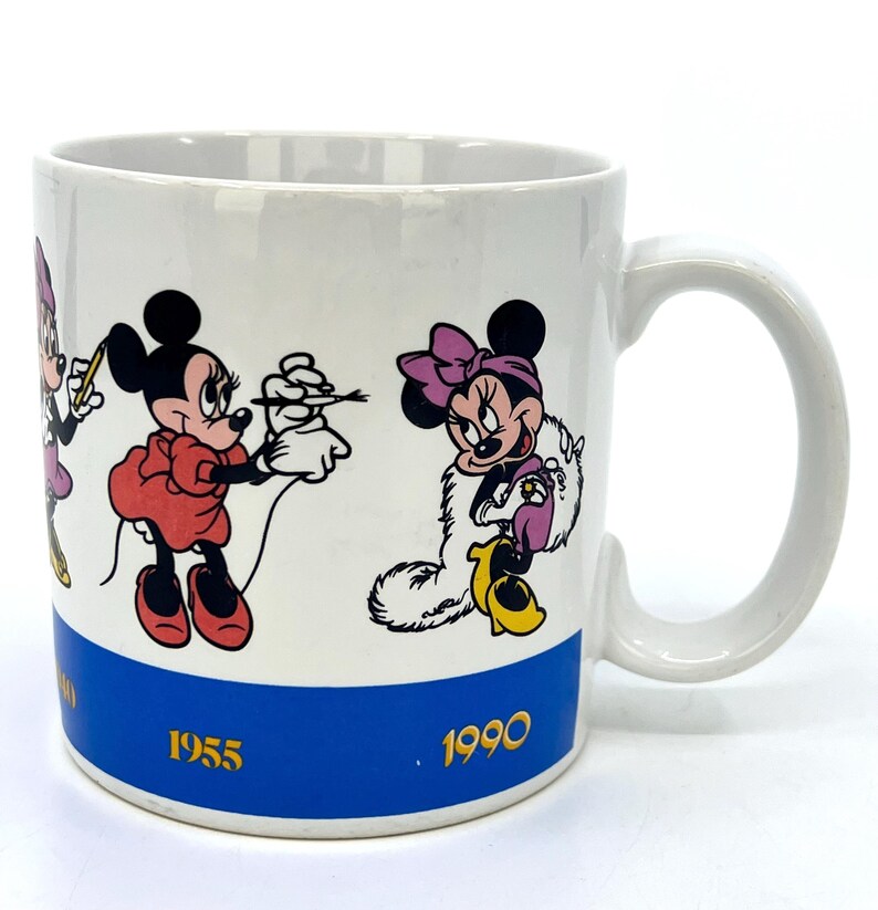 Op de afbeelding: Witte keramische mok met Minnie Mouse in verschillende poses. De mok heeft een blauwe band aan de onderkant met de jaren 1955 en 1990 in goud. De handgreep is wit en de mok is cilindrisch.