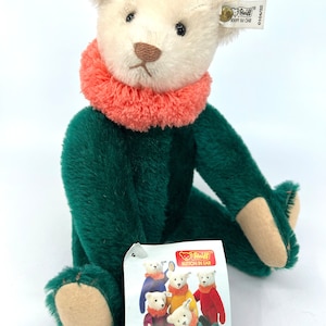 May include: A small, white plush teddy bear wearing a green outfit and a pink collar. The bear is sitting with its legs crossed and has a button in its ear. The bear is labeled "Steiff Button in Ear" and "Circus Dolly Bear".