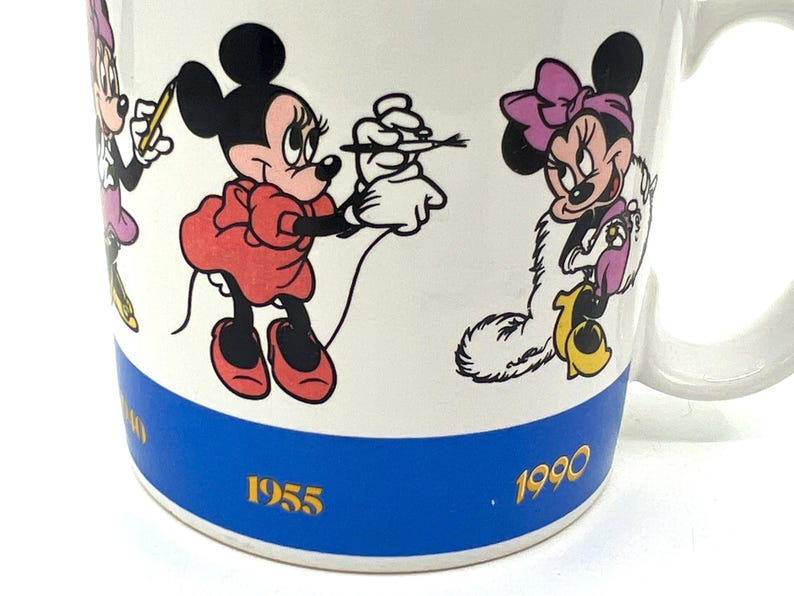 Op de afbeelding: Witte keramische mok met illustraties van Minnie Mouse in verschillende poses. De mok heeft een blauwe band met de jaren 1955 en 1990. De handgreep is wit.