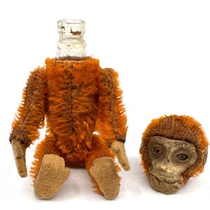 Op de afbeelding: Een vintage pluche aap speelgoed met een glazen fles als hoofd. De aap is bruin en oranje met een jute-achtige textuur. De aap mist zijn hoofd en de glazen fles is leeg.