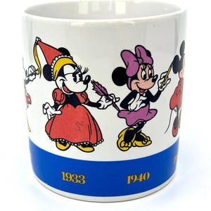 Op de afbeelding: Witte keramische mok met illustraties van Minnie Mouse in verschillende outfits. De mok heeft een blauwe band aan de onderkant met de jaren 1933 en 1940 in goud gedrukt. De stripfiguren zijn in heldere kleuren.