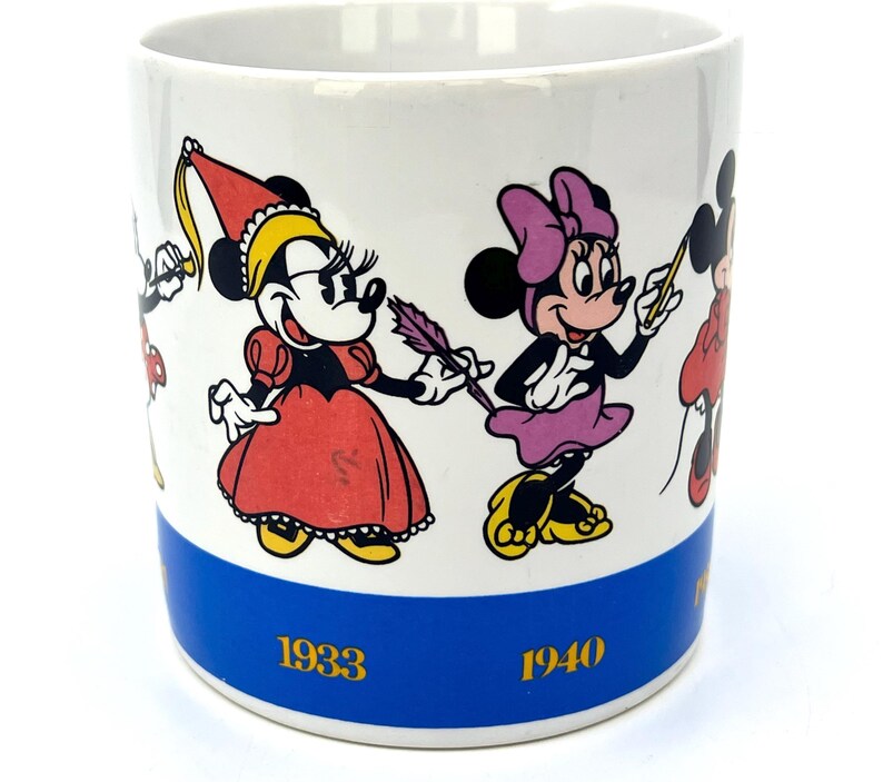 Op de afbeelding: Witte keramische mok met illustraties van Minnie Mouse in verschillende outfits. De mok heeft een blauwe band aan de onderkant met de jaren 1933 en 1940 in goud gedrukt. De stripfiguren zijn in heldere kleuren.