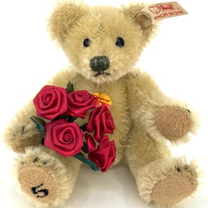May include: A vintage Steiff teddy bear with a bouquet of red roses. The plush bear has a light tan fur, black eyes, and a green stitched nose. A small tag is attached to the ear. The bear is holding a bouquet of red roses.