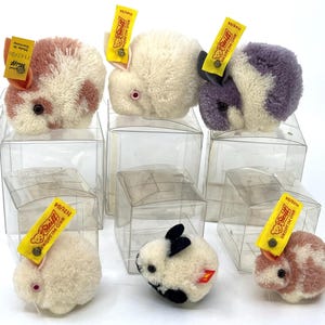 Puede incluir: Una colección de pequeños conejitos de peluche en cajas de plástico transparente. Los conejitos son blancos, rosas y morados, con detalles en negro o rosa. Cada conejito tiene una etiqueta amarilla con el logotipo de Steiff y texto.