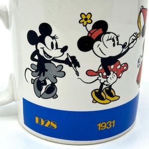 Op de afbeelding: Witte keramische mok met illustraties van Mickey en Minnie Mouse. Mickey is afgebeeld in zwart en grijs, met een pen. Minnie draagt een rode jurk met stippen. De mok heeft een blauwe band met de jaren 1928 en 1931.