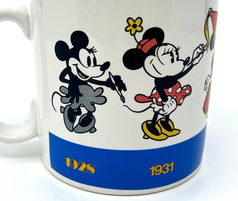 Op de afbeelding: Witte keramische mok met illustraties van Mickey en Minnie Mouse. Mickey is afgebeeld in zwart en grijs, met een pen. Minnie draagt een rode jurk met stippen. De mok heeft een blauwe band met de jaren 1928 en 1931.