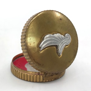 May include: A gold metal compact with a silver winged design on the lid. The compact is open and the inside is lined with a red and white fabric.